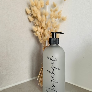 May include: A frosted glass pump bottle with a black pump top and a black label that says "Duschgel". The bottle is sitting on a counter next to a bouquet of dried pampas grass.
