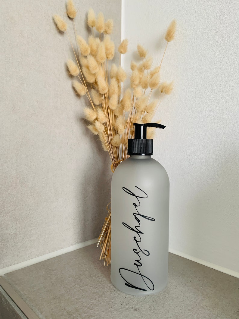 May include: A frosted glass pump bottle with a black pump top and a black label that says "Duschgel". The bottle is sitting on a counter next to a bouquet of dried pampas grass.