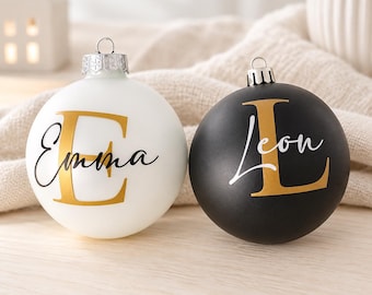 May include: Two round Christmas ornaments, one matte white and one matte black, each featuring a large gold serif letter and a name in black cursive script. The white ornament displays the letter E and the name Emma, while the black ornament shows the letter L and the name Leon. Both have silver metal caps.