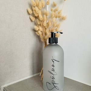 May include: A frosted gray glass pump bottle with a black pump top. The bottle is labeled with the word "Sp&uuml;lung" in black cursive writing. The bottle is sitting on a gray tiled surface with a bouquet of dried beige pampas grass behind it.