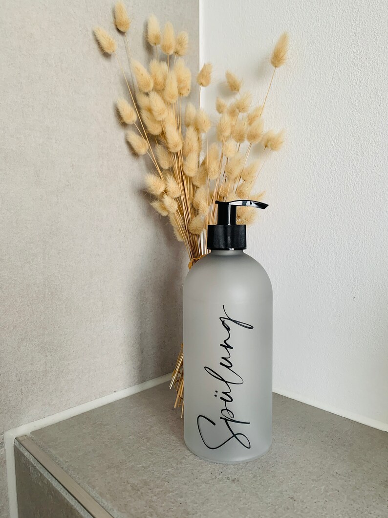 May include: A frosted gray glass pump bottle with a black pump top. The bottle is labeled with the word "Sp&uuml;lung" in black cursive writing. The bottle is sitting on a gray tiled surface with a bouquet of dried beige pampas grass behind it.