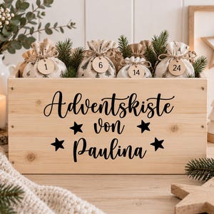 Könnte beinhalten: Eine rechteckige Holzkiste in heller Farbe mit dem schwarzen Schriftzug Adventskiste von Paulina und vier Sternsymbolen. Darin befinden sich mehrere kleine Stoffbeutel mit Kordelzug und Baummustern, die jeweils ein rundes Holzetikett mit Zahlen tragen.