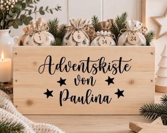 Könnte beinhalten: Eine rechteckige Holzkiste in heller Farbe mit dem schwarzen Schriftzug Adventskiste von Paulina und vier Sternsymbolen. Darin befinden sich mehrere kleine Stoffbeutel mit Kordelzug und Baummustern, die jeweils ein rundes Holzetikett mit Zahlen tragen.