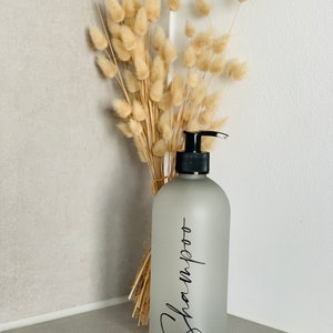 May include: A frosted glass bottle with a black pump top and a black label that says "Shampoo". The bottle is sitting on a white shelf with a bouquet of dried pampas grass behind it.