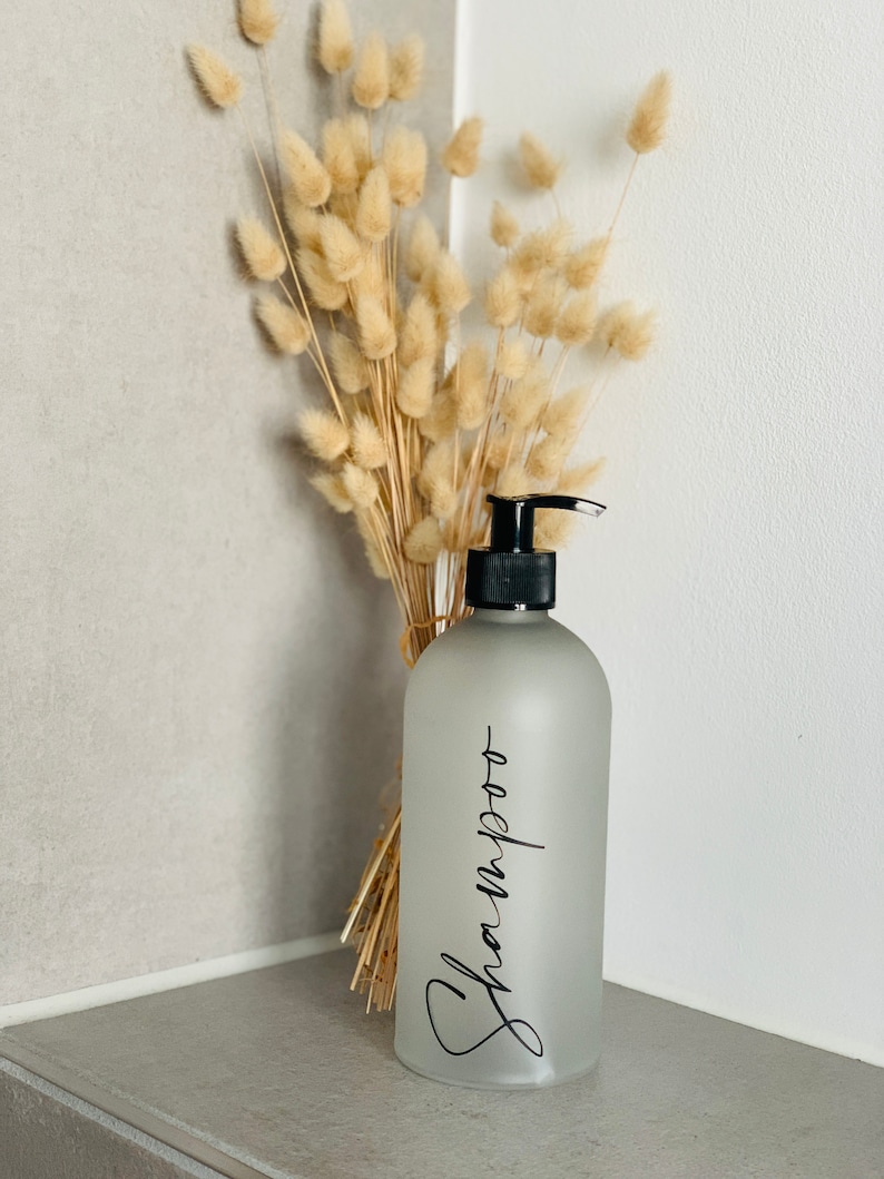 May include: A frosted glass bottle with a black pump top and a black label that says "Shampoo". The bottle is sitting on a white shelf with a bouquet of dried pampas grass behind it.