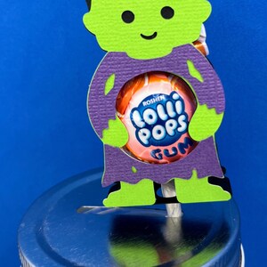 May include: A paper cutout of a green-skinned character with black hair and a purple shirt, holding a round Roshen Lolli Pops Gum lollipop. The cutout is attached to the silver metal lid of a glass jar, featuring a small slot on the lid surface.
