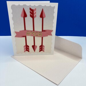 May include: A greeting card with three red arrows and the word "AMOR" on a pink banner. The card is inside a matching envelope, perfect for Valentine's Day or expressing love. The card has a decorative border.