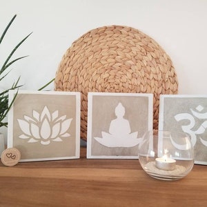 Könnte beinhalten: Vier quadratische Beton-Wanddekorationen mit einer weißen Lotusblume, einem meditierenden Buddha, dem Om-Symbol und einem Kerzenhalter. Die Stücke sind auf einer Holzoberfläche mit einer geflochtenen Strohmatte dahinter angeordnet.