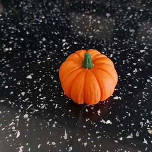May include: A small, orange pumpkin with a green stem. The pumpkin has a ribbed texture, likely a decorative item for autumn or Halloween. It sits on a black speckled surface. A seasonal decoration.