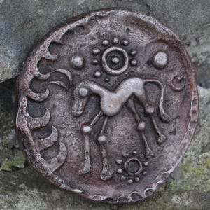 Peut inclure: Médaillon rond de couleur bronze vieilli avec un motif en relief. Le motif représente un cheval stylisé au centre, entouré de formes solaires et lunaires. Le médaillon a une surface rugueuse et texturée.