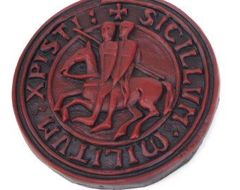 May include: A circular, red emblem with a black border. The emblem features two knights on a horse, a cross, and Latin text. The design is reminiscent of a historical seal or coin, with a weathered appearance.