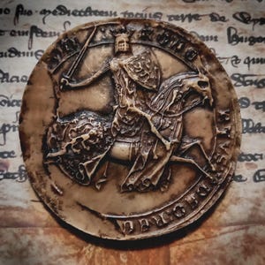 Puede incluir: Una escultura en relieve circular de estilo antiguo, en tonos beige y marrón. Representa a un caballero a caballo, empuñando una espada y un escudo. El caballero está acorazado y lleva un casco. La escultura tiene un aspecto envejecido y detallado, con texto alrededor del perímetro.