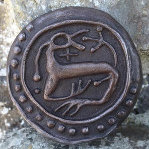 May include: A circular, bronze-coloured metal pendant with a raised design of a stylised deer with antlers. The deer is surrounded by a circle of small dots.