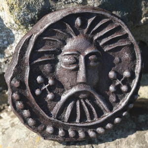 May include: A weathered, circular, bronze-colored relief sculpture of a face with a beard and sun-like rays emanating from the head. The face has closed eyes and is surrounded by a beaded border and decorative elements.
