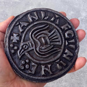 May include: A round, dark grey metal plaque with a stylised bird design and the inscription "F.N.I.V.E.G.I.N.I.K." around the edge.