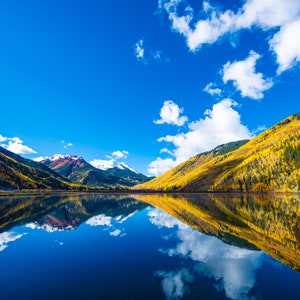 May include: A mountain lake with a reflection of the surrounding fall foliage and clouds in the water. The mountains are covered in trees with yellow, orange, and red leaves. The sky is blue with white clouds.