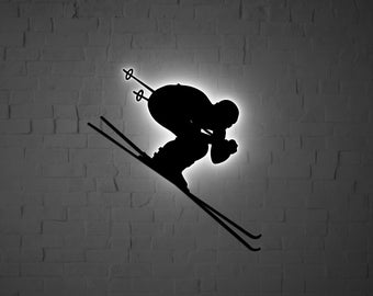 Op de afbeelding: Silhouet van een skiër in de lucht, met uitgespreide ski's, tegen een achtergrond van een witte bakstenen muur.