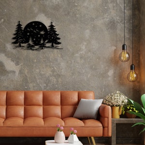 K&ouml;nnte beinhalten: Schwarze Wandkunst aus Metall, die einen Vollmond mit drei Kiefern und Schneeflocken darstellt. Die Kunst h&auml;ngt an einer grauen Betonwand &uuml;ber einem braunen Ledersofa.