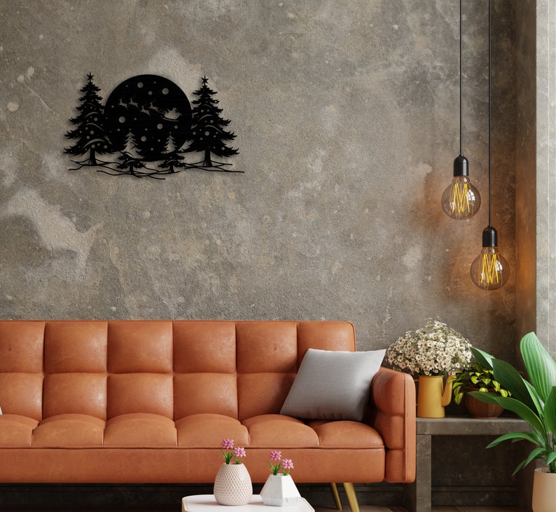 K&ouml;nnte beinhalten: Schwarze Wandkunst aus Metall, die einen Vollmond mit drei Kiefern und Schneeflocken darstellt. Die Kunst h&auml;ngt an einer grauen Betonwand &uuml;ber einem braunen Ledersofa.
