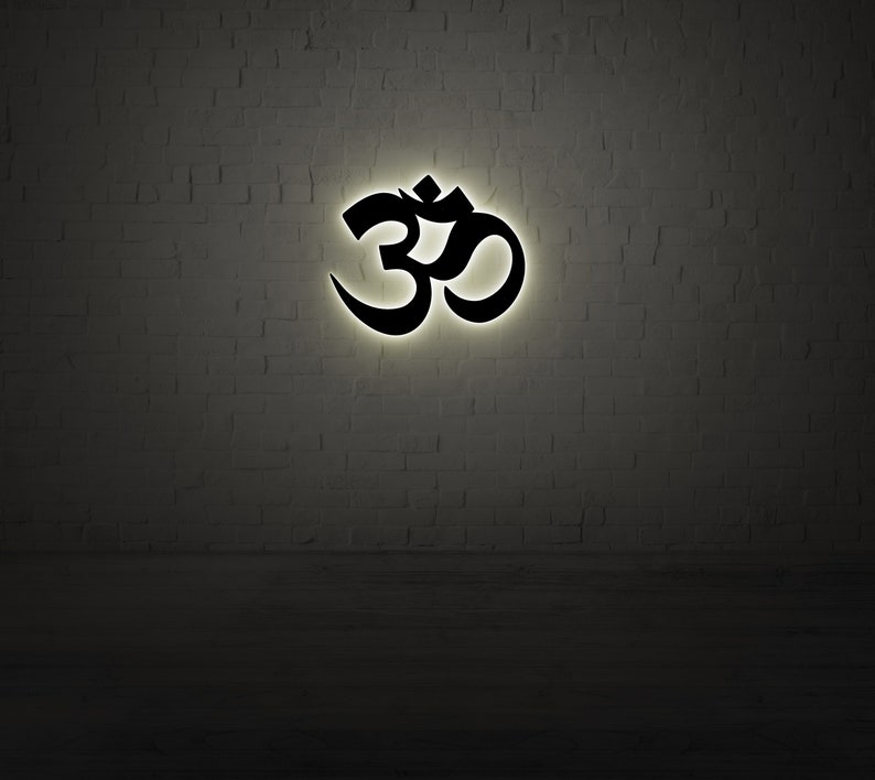 May include: A black Om symbol illuminated against a white brick wall.