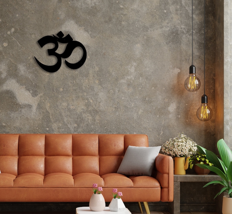 May include: A brown leather sofa with a grey throw pillow and two small potted plants with pink flowers. The wall behind the sofa is a textured grey and features a black metal Om symbol.