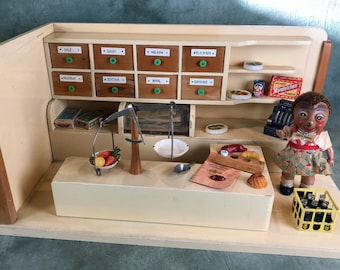 May include: A vintage wooden toy shop playset with a doll shopkeeper, shelves, a scale, and various toy food items. The shop has a white counter and a brown wooden frame.
