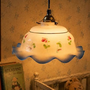 Puede incluir: Una lámpara colgante con una pantalla de vidrio blanco esmerilado que presenta un diseño floral pintado a mano y un borde ondulado con tonos azules. La lámpara está suspendida por un soporte de metal oscuro y emite una luz cálida.