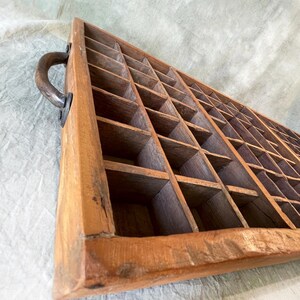 May include: A vintage wooden printer's tray with numerous small compartments. The tray is rectangular with a handle on one side. The wood has a warm, natural tone with visible grain and some signs of wear, suggesting age and use.