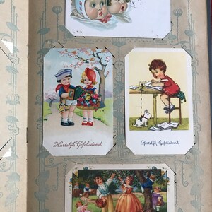 May include: A vintage scrapbook with four illustrated cards featuring children. The first card shows two children looking up at a spider. The second card shows two children, one playing an accordion and the other holding flowers. The third card shows a child writing at a desk with a white rabbit nearby. The fourth card shows children picking cherries from a tree. The text on the cards reads "Hartelijk Gefeliciteerd" and "Le temps des cerises."