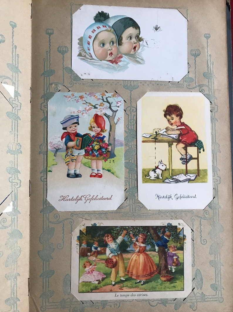 May include: A vintage scrapbook with four illustrated cards featuring children. The first card shows two children looking up at a spider. The second card shows two children, one playing an accordion and the other holding flowers. The third card shows a child writing at a desk with a white rabbit nearby. The fourth card shows children picking cherries from a tree. The text on the cards reads "Hartelijk Gefeliciteerd" and "Le temps des cerises."