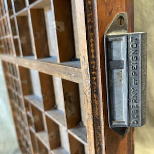 May include: A vintage wooden printer's type drawer featuring a grid of small compartments for organizing items. A rectangular metal handle is attached to the side, clearly embossed with the text DEBERNY ET PEIGNOT in capital letters.