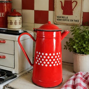 Puede incluir: Una cafetera roja de esmalte con lunares blancos, un borde negro y un asa curva. La cafetera forma parte de una escena de cocina con un fondo de baldosas rojas y blancas y un letrero que dice "KOFFIE".