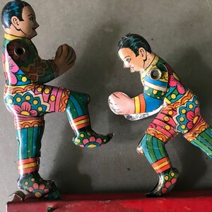 May include: Two colorful tin toy figures of boxers, one in a blue and green outfit and the other in a pink and green outfit, are posed in a fighting stance. The figures are attached to a red metal base.