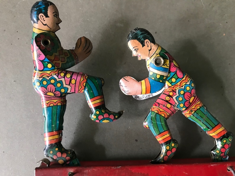 May include: Two colorful tin toy figures of boxers, one in a blue and green outfit and the other in a pink and green outfit, are posed in a fighting stance. The figures are attached to a red metal base.