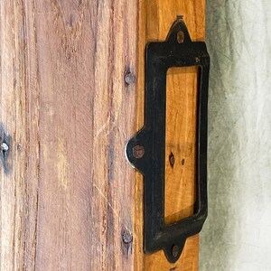 May include: Close-up of a wooden plank with a rectangular black metal label holder attached. The wood has a natural grain and is secured with small nails. The metal holder has rounded edges.