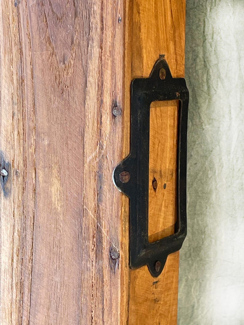 May include: Close-up of a wooden plank with a rectangular black metal label holder attached. The wood has a natural grain and is secured with small nails. The metal holder has rounded edges.