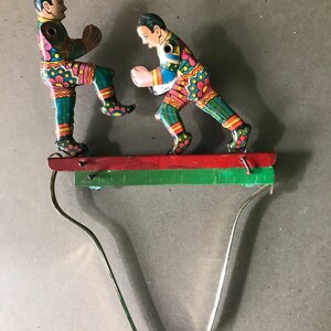 May include: Two colorful tin toy figures of boxers, wearing red, green, blue, and yellow patterned outfits, are posed in a fighting stance on a red and green platform. The figures are attached to a metal spring.
