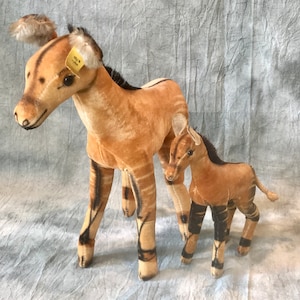 May include: Two plush Okapi toys, one larger and one smaller, standing side-by-side. The larger Okapi is brown with black stripes and has a yellow tag on its ear. The smaller Okapi is also brown with black stripes.