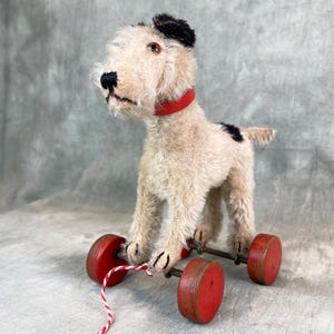 Puede incluir: Un perro de peluche vintage blanco y negro con un collar rojo y orejas negras. El perro está en un carro de juguete de madera con ruedas rojas y una cuerda roja y blanca.