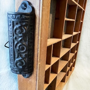 May include: A vintage wooden printer's tray with multiple small compartments. A black, ornate match holder is mounted on the side. The wood has a warm, aged tone, and the tray is empty.