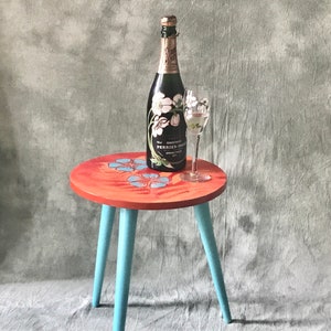 Puede incluir: Una mesa pequeña de tres patas con una tapa roja pintada con diseños florales azules. Una botella de champán Perrier-Jouët y una copa están sobre la mesa.