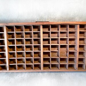 May include: A rectangular wooden printer's tray with numerous small, square compartments. The tray is made of light brown wood and has a rustic, aged appearance. It features two small handles on the sides.