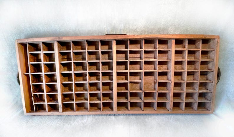 May include: A rectangular wooden printer's tray with numerous small, square compartments. The tray is made of light brown wood and has a rustic, aged appearance. It features two small handles on the sides.
