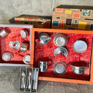May include: A vintage toy kitchen set with miniature pots, pans, and utensils. The set includes a variety of metal cookware with red handles and lids, displayed in a framed arrangement with a red patterned background. Boxes with colorful designs are in the background.