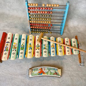 May include: A vintage children's toy set featuring a blue abacus with wooden beads, a colorful xylophone with a wooden mallet, and a small harmonica with a painted scene. The xylophone has illustrations of cartoon characters.