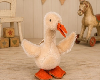 Puede incluir: Un juguete de peluche con forma de ganso de suave pelaje color crema, que presenta un pico de fieltro naranja brillante y patas palmeadas a juego. El ganso se muestra en posición erguida con las alas extendidas y costuras visibles.