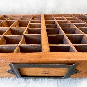 May include: A vintage wooden printer's tray with numerous small compartments. The tray is made of light brown wood and features a dark metal handle. The compartments are arranged in a grid pattern, ideal for organizing small items.