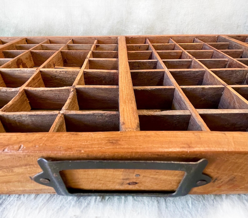 May include: A vintage wooden printer's tray with numerous small compartments. The tray is made of light brown wood and features a dark metal handle. The compartments are arranged in a grid pattern, ideal for organizing small items.