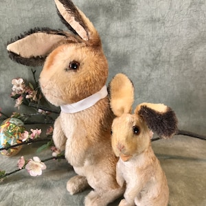 May include: Two plush bunny rabbits, one larger than the other, are standing side-by-side. The larger bunny is brown and tan with a white ribbon around its neck. The smaller bunny is tan with brown ears. Both bunnies have black eyes and are made of soft, fluffy material.
