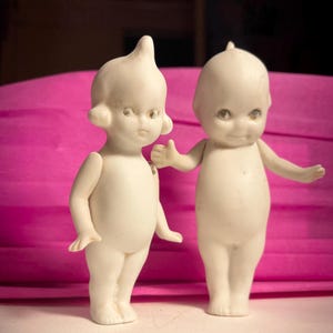 May include: Two antique bisque dolls with a cream-colored finish. The dolls have detailed facial features, including small eyes and delicate limbs. One doll has a small tuft of hair, while the other has a small point on top of its head. They stand on a white surface against a pink backdrop.